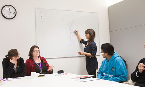 Landscape image of a room with three people sat at a table and a fourth writing on a whiteboard
