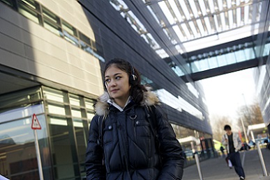 A long, dark haired student walks between buildings wearing large headphones with hands in pockets.