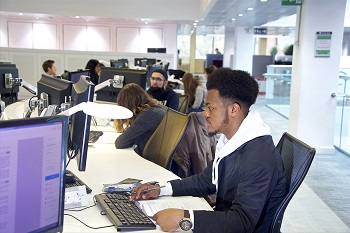 A student sits at a library desk looking at a computer screen. Other students can be seen in the background.