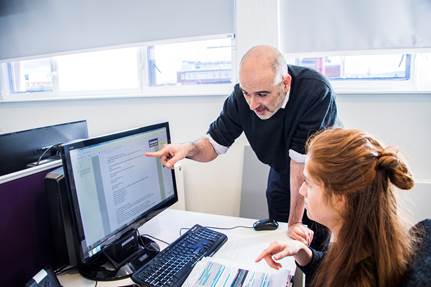 A woman works at a computer while the man stood next to her points to the document on-screen.