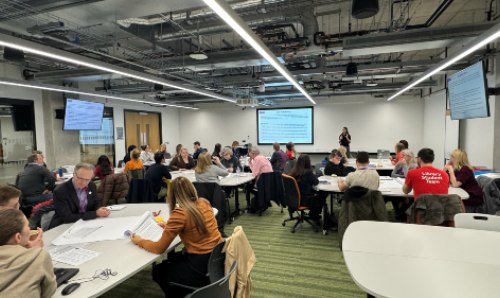 Image from the workshop with staff sat round tables, Bliss Ross presenting