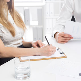 Two members of staff sat at a desk, one reading from a page, the other writing down 