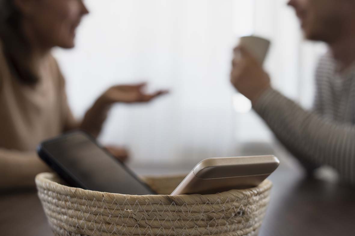 Mobile phones are placed in a basket at the front of the image whilst a man and a woman have a discussion in the background