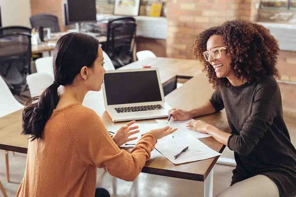 Colleagues engaged in a coaching discussion around a desk in an office. There is a laptop on the table and the two colleagues are smiling, enjoying their conversation