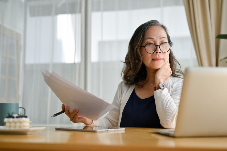 Concentrated asian middle aged female teacher or businesswoman in glasses sitting at desk using portable computer and examining paperwork