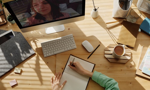 ariel view, person at a busy desk writing in a notebook, while the computer in front of them shows a video call in progress