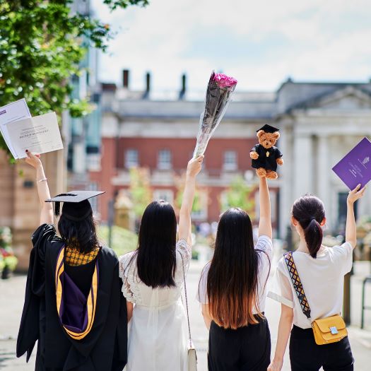 4 students with backs to camera. Left one is wearing graduation gown and other 3 are in white tops and black bottoms. All lifting up various objects, degree certificate, flowers, bear and  programme.