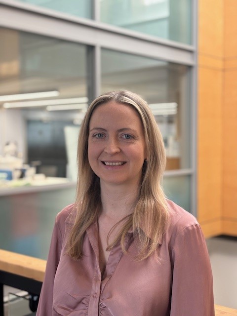academic woman with blonde hair stood in an office building smiling at the camera