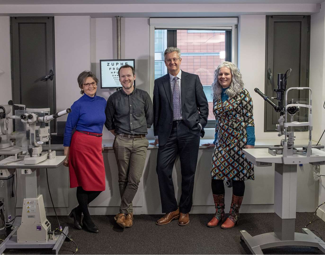 A woman, a man, a man and a woman stand next to each other in an optometry lab, smiling at the camera.