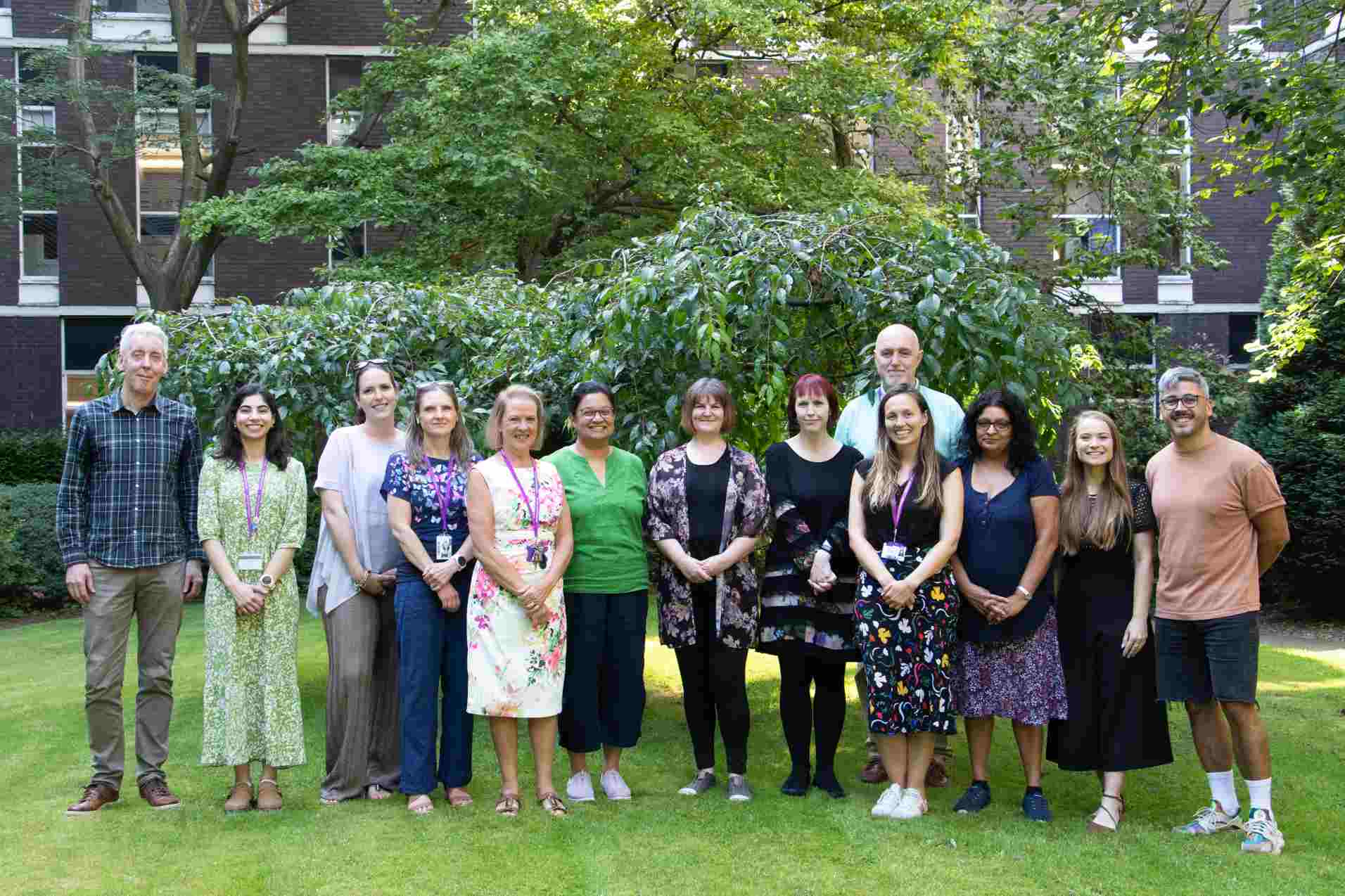 All 13 members of the core team standing smiling in a line outside on a lawn in front of some leafy shrubs