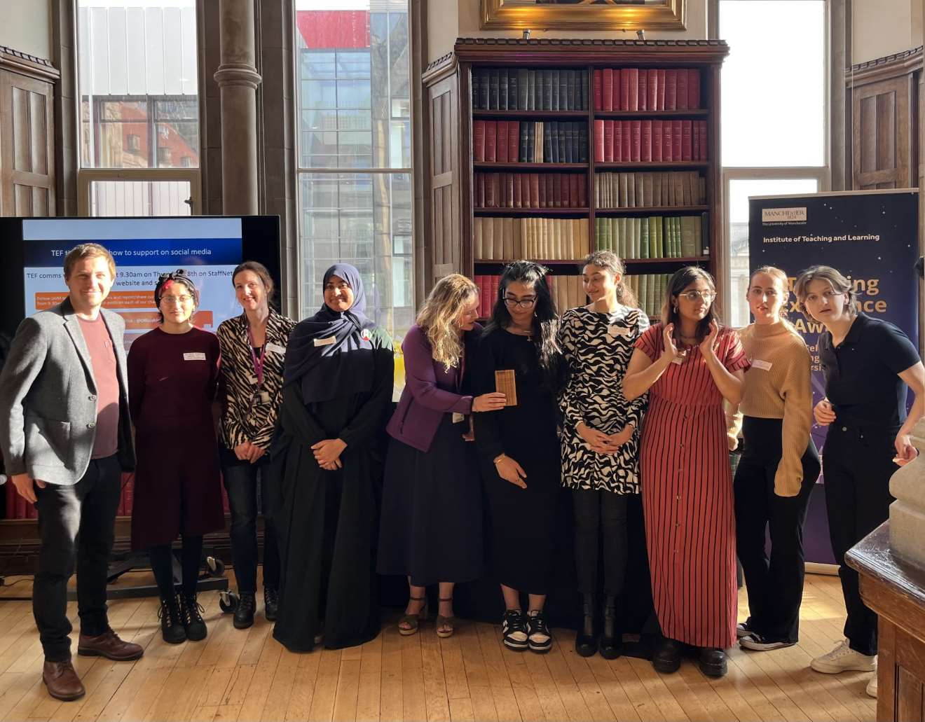 A group of staff and students standing in front of a tall window and tall built-in wooden bookcase in the university of Christie Bistro (the old library) 