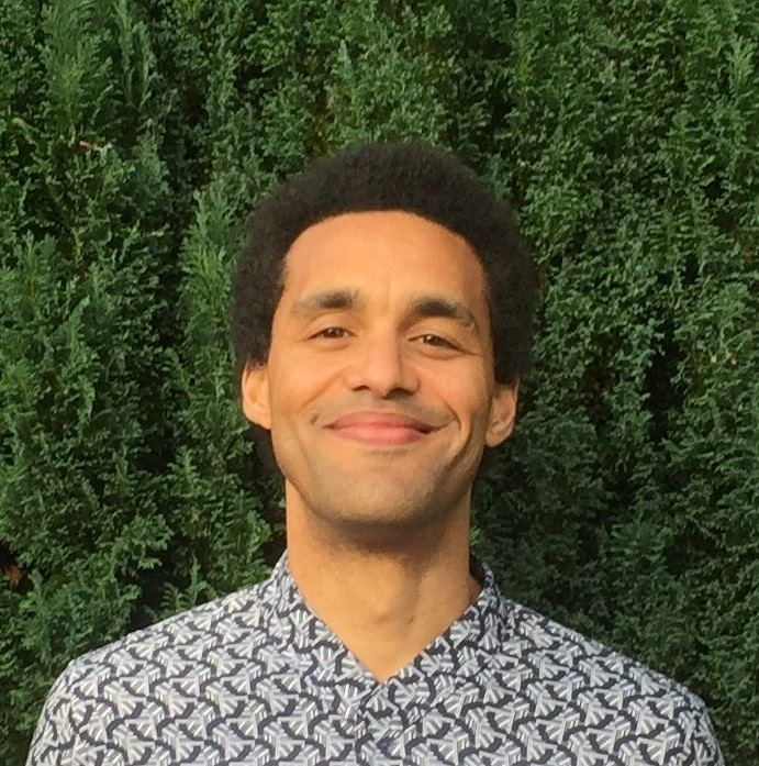 Photograph of Adam Danquah smiling in front of a green hedge