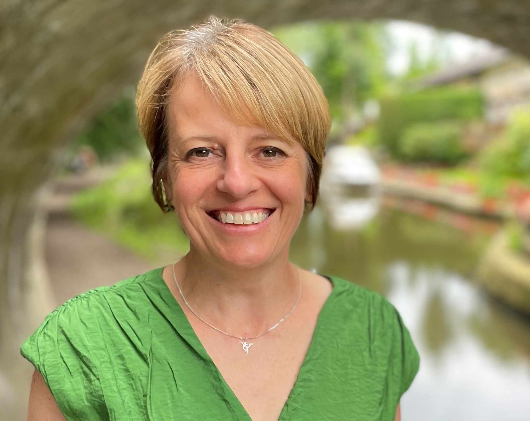 Photo of Ang Davies, a woman with short blonde hair wearing a green top and smiling in front of a canal.