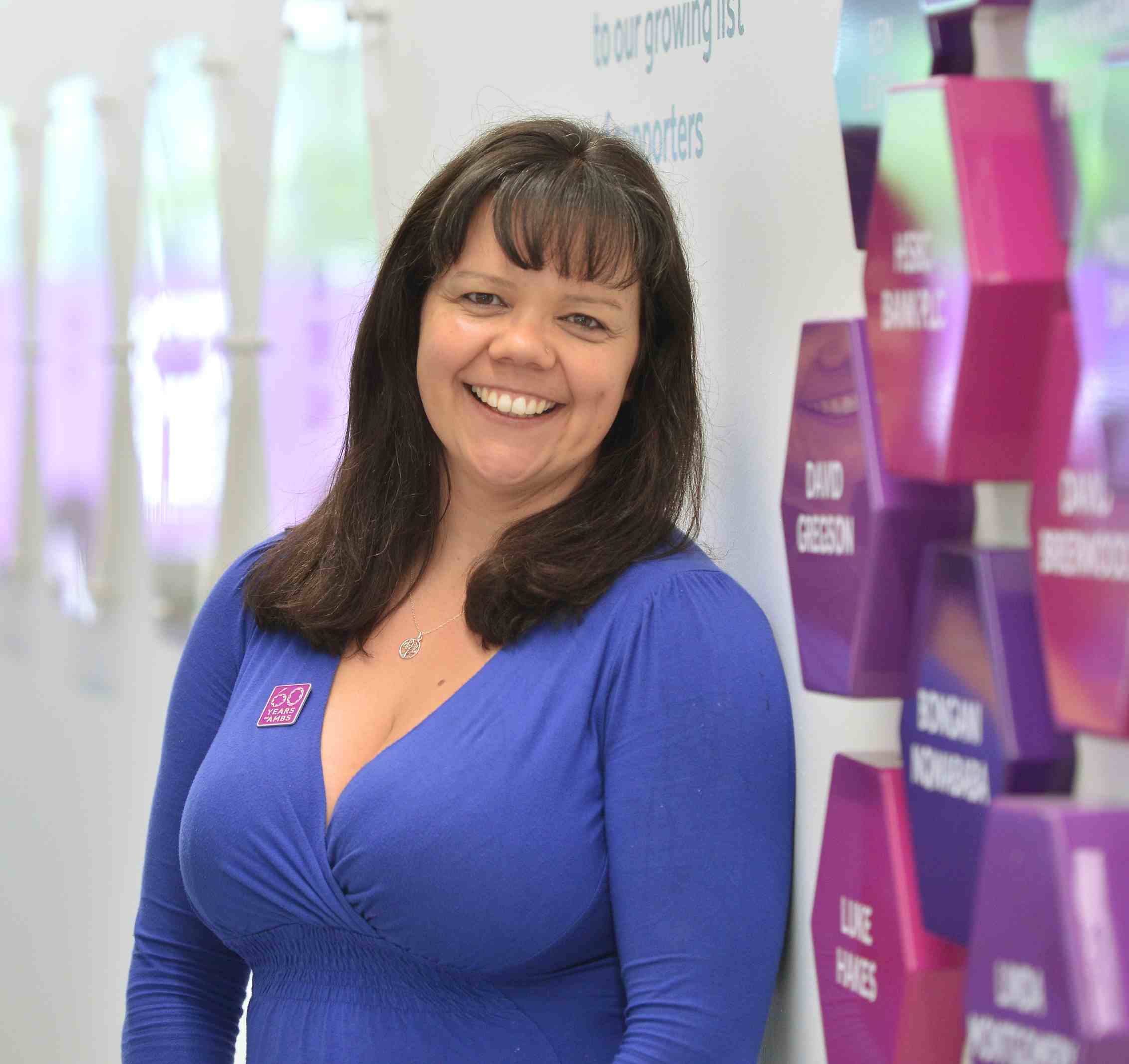 Photo of Jenni Rose, a woman with long dark hair wearing a purple top, smiling in front of the donor wall in the Alliance Manchester Business School building.